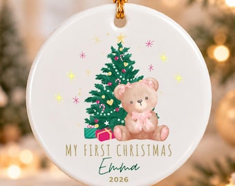 Puede incluir: Un adorno navideño redondo de cerámica blanca con un árbol de Navidad verde, un oso de peluche marrón con un lazo rosa y regalos. El texto dice MY FIRST CHRISTMAS, el nombre Emma y el año 2026 en color dorado y verde oscuro.