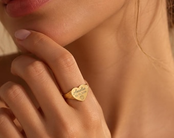 Puede incluir: Un anillo dorado en forma de corazón con el nombre Amelia grabado, la fecha 02.10.2026 y un pequeño icono de huella. El anillo tiene un acabado metálico pulido y se lleva en un dedo, mostrando el detalle del grabado personalizado.