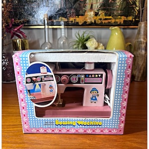 May include: A pink battery-operated sewing machine toy inside a cardboard box with a blue and white checkered border. The machine features a graphic of a child in a blue hat and dress, a white spool, and a black control panel with geometric shapes.