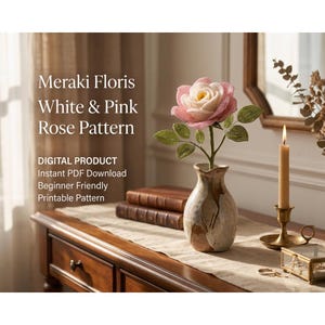 May include: A digital product listing for a Meraki Floris White & Pink Rose Pattern. The image features a handcrafted pink and white rose with green leaves in a ceramic vase, next to a lit candle and books, with text detailing the PDF download.