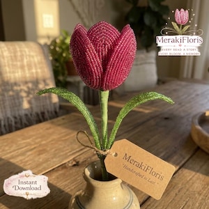 May include: A deep magenta beaded tulip flower with green beaded leaves and a stem, standing in a small ceramic vase. A tan rectangular paper tag is tied to the stem with twine, featuring the printed text MerakiFloris Handcrafted to Last.