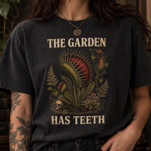 Puede incluir: Una camiseta de manga corta gris carbón con un estampado gráfico de una venus atrapamoscas, una planta jarra y una pequeña calavera. El texto THE GARDEN HAS TEETH está impreso en una fuente beige sobre y debajo de la ilustración.