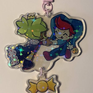 May include: A clear acrylic keychain featuring a holographic star pattern over an illustration of two cartoon characters holding hands, one with lime green hair and a crown, the other with red hair, plus a small yellow candy-shaped charm attached.