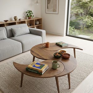 May include: A two-piece wooden coffee table set with a curved, interlocking design. The tables feature a natural wood grain finish and tapered legs. On the surface are three small bowls containing nuts, two ceramic mugs, and two books titled "Yasam Sanati" and "Renklerin Gizemi."