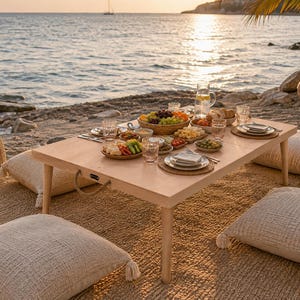 May include: A low-profile rectangular wooden picnic table with rope handles, set with various plates of fruit, cheese, and snacks, surrounded by four square textured beige floor cushions with tassels on a woven rug near the ocean shore.