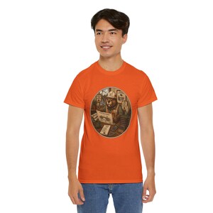 Könnte beinhalten: Ein orangefarbenes Kurzarm-T-Shirt mit einer kreisförmigen Grafik eines Bibers, der einen Schutzhelm und eine Warnweste trägt und an einem Schreibtisch mit Bauplänen, einem Taschenrechner und verschiedenen Büroartikeln sitzt.