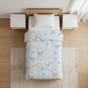 Op de afbeelding: Een lichtblauw dekbed met een herhalend patroon van witte stripfiguren met lange oren, roze en blauwe hartjes, strikjes en sterretjes. De beddengoedset bevat een bijpassende witte kussensloop op een bedframe met twee witte nachtkastjes.