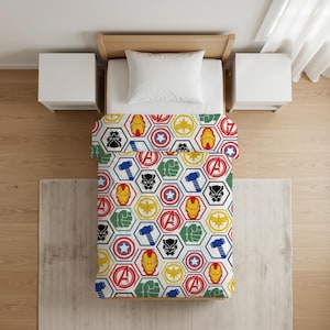 Puede incluir: Una cama individual con un edredón blanco que presenta un patrón hexagonal repetitivo. Cada hexágono muestra iconos coloridos de símbolos de superhéroes, incluyendo máscaras, escudos, martillos y puños en colores rojo, azul, amarillo, verde y negro.