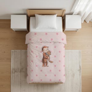 Op de afbeelding: Een lichtroze dekbedovertrek met een herhalend patroon van kleine roze sterren en een centrale illustratie van een bruine aap die een kleine beige kat vasthoudt. Het beddengoed ligt op een houten bedframe met een wit kussen en twee witte nachtkastjes.