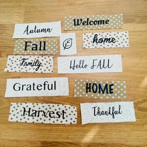 May include: A collection of rectangular fabric strips featuring beige and white patterns with black printed text including Autumn, Welcome, Fall, home, Family, Hello FALL, Grateful, HOME, Harvest, and Thankful, plus a small square with a leaf icon.