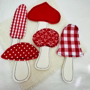 May include: Four fabric mushroom cutouts with cream-colored stems and red caps featuring various patterns including red and white gingham, solid red, white polka dots on red, and a white floral print on red, all outlined with black stitching.