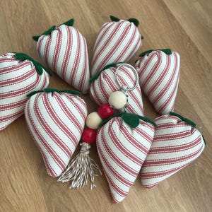 May include: A collection of fabric strawberry-shaped ornaments featuring a red and white striped pattern with green felt leaves at the top. One strawberry is attached to a silver metal keychain with red and white wooden beads and a tassel.