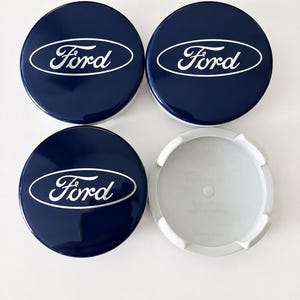 May include: A set of four circular wheel centre caps featuring a glossy dark blue finish with the white Ford logo centred on the front. The caps have a smooth, rounded top surface and a light grey plastic underside with moulded mounting clips.