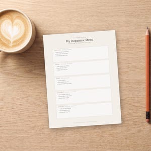 May include: A white paper sheet titled My Dopamine Menu with sections for starters, mains, sides, desserts, and specials. The sheet is positioned next to a ceramic mug filled with coffee featuring heart-shaped latte art and an orange pencil.