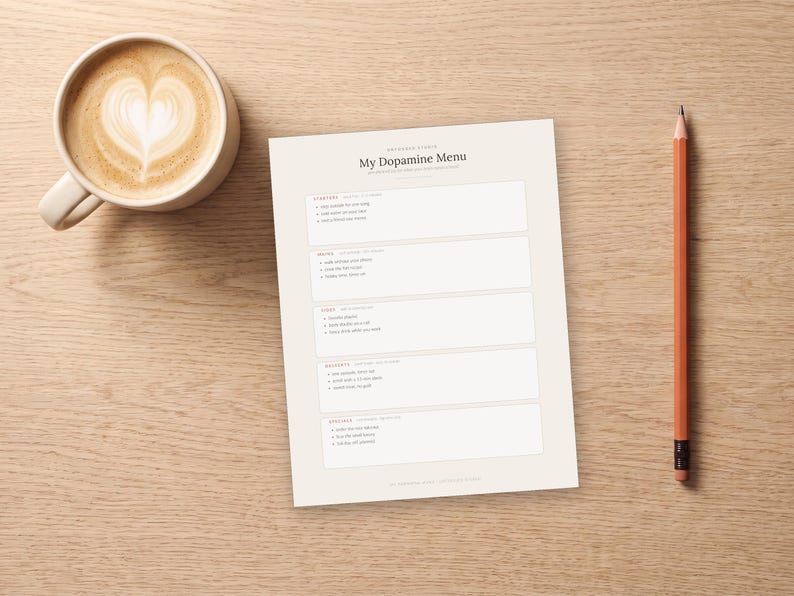 May include: A white paper sheet titled My Dopamine Menu with sections for starters, mains, sides, desserts, and specials. The sheet is positioned next to a ceramic mug filled with coffee featuring heart-shaped latte art and an orange pencil.