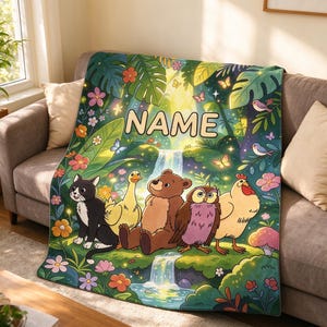 Op de afbeelding: Een rechthoekige deken met een kleurrijke jungle-scène, een waterval en de tekst NAME. De illustratie toont een zwart-witte kat, een gele eend, een bruine beer, een paarse uil en een kip, omringd door diverse bloemen en planten.