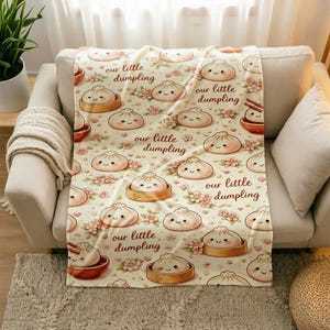 May include: A cream-coloured throw blanket featuring a repeating pattern of cartoon dumplings with smiling faces, some in bamboo steamers or bowls, interspersed with small pink flowers and the repeating text our little dumpling.