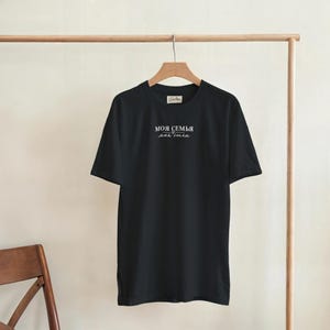 Könnte beinhalten: Ein schwarzes kurzärmeliges T-Shirt mit weißem Text auf der Brust, der "МОЯ СЕМЬЯ моя сила" lautet, mit einem kleinen Herzsymbol zwischen den Zeilen. Das Kleidungsstück hängt auf einem Holzkleiderbügel vor einem hellen, einfarbigen Hintergrund.