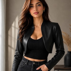 Peut inclure: Une veste courte noire à manches longues avec une finition lisse, portée sur un débardeur noir côtelé et un jean noir. La veste présente un design sans col avec une ouverture frontale incurvée qui s'arrête au-dessus de la taille.