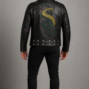 Op de afbeelding: Een zwart leren jack met een grote geborduurde slangafbeelding op de rug in groen- en geeltinten. Het jack is voorzien van zilverkleurige metalen studs langs de kraag en de onderzoom, en zilveren ritsen op de mouwen.