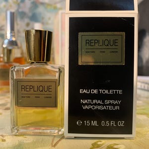 May include: A clear glass perfume bottle with a gold cap and a label reading REPLIQUE NEW YORK PARIS LONDON, next to a black and white rectangular box with the same text, EAU DE TOILETTE, NATURAL SPRAY, and the volume 15 ML 0.5 FL OZ.