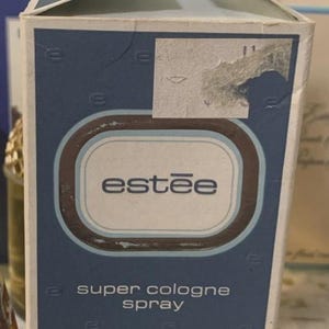 May include: A rectangular Estée Lauder super cologne spray box in blue and white. The front features the text 'estée', 'super cologne spray', and 'estée lauder 1.85 FL. OZ.' with a repeating 'e' logo pattern. The top flap shows repeated brand text.