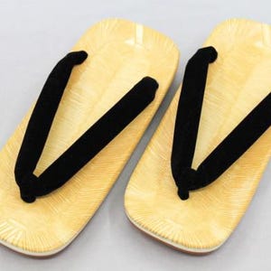 May include: A pair of zori sandals featuring light yellow, textured footbeds with a radiating line pattern and thick, black velvet-like thong straps. The rectangular-shaped soles have rounded edges and a layered construction with a brown base.