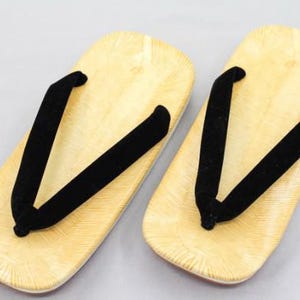May include: A pair of zori sandals featuring light tan, wood-grain patterned footbeds and thick, black velvet thong straps. The sandals have a flat, rectangular base with rounded edges, showcasing a textured surface design on the footbed.