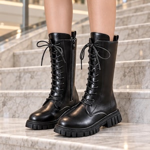 Könnte beinhalten: Ein Paar schwarze wadenhohe Stiefel mit Schnürung vorne, passenden schwarzen Schnürsenkeln, seitlichem Reißverschluss, abgerundeter Zehenpartie und einer dicken, profilierten Sohle. Die Stiefel haben eine glatte Oberfläche und eine Zugschlaufe.