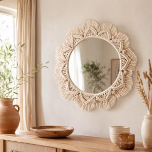 May include: A round wall mirror framed by intricate off-white macrame cord woven into a sunburst pattern with knotted details and a fringed edge. The mirror features a circular glass center reflecting a room interior with a green plant.