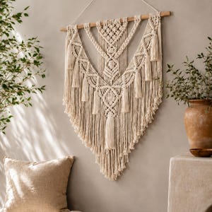 Puede incluir: Un gran tapiz de macramé en color blanco roto con intrincados patrones de nudos, diseños geométricos y múltiples borlas colgantes. La pieza está suspendida de un listón de madera horizontal con un cordón blanco fino para colgar.