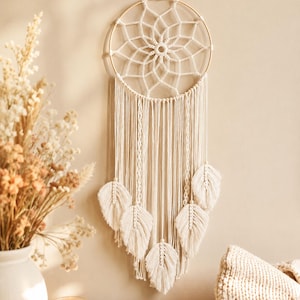 Op de afbeelding: Een crèmekleurige macramé wandhanger met een ronde houten ring met een ingewikkeld geweven webpatroon in het midden. Vijf macramé veren hangen aan de onderkant van de ring, opgehangen aan lange, dunne, geknoopte katoenen koorden.