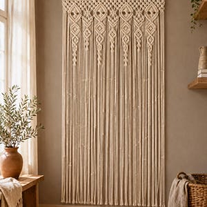 May include: A large off-white macrame wall hanging suspended from a wooden rod. The piece features an intricate knotted pattern at the top with diamond-shaped motifs, transitioning into long, vertical hanging cords that reach the floor.