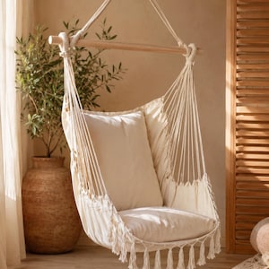 May include: A hanging chair made of off-white fabric with a wooden spreader bar and thick rope suspension. The seat features a matching back cushion and a seat cushion, finished with decorative fringe tassels along the bottom edge of the fabric.