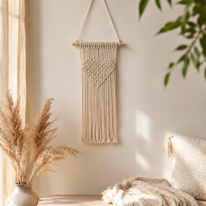 May include: A cream-colored macrame wall hanging featuring a central heart pattern created with intricate knots. The textile piece is suspended from a horizontal wooden dowel by a single cord, with long, straight fringe hanging from the bottom edge.