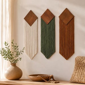Op de afbeelding: Drie wandkleden met ruitvormige donkerhouten bovenkanten en lange, rechte vezelfranjes in de kleuren crème, olijfgroen en terracotta. Elk stuk is verticaal gemonteerd en toont een strak geometrisch ontwerp met trapsgewijs vallende textielstrengen.