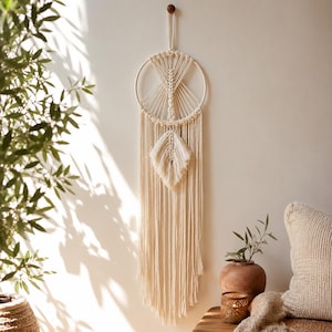 May include: A cream-colored macrame wall hanging featuring a circular frame with intricate knotted patterns and a central leaf-shaped element. Long, flowing fringe extends from the bottom of the hoop, creating a vertical, textured design.