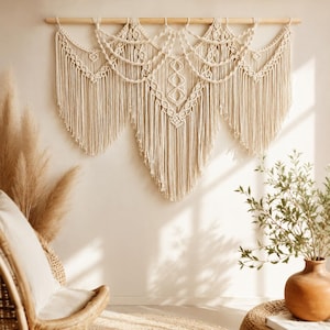 May include: A large off-white macrame wall hanging suspended from a horizontal wooden dowel. The piece features intricate knotted patterns, including diamond shapes and draped swags, finished with long, flowing fringe that forms a pointed shape.