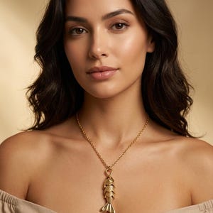 May include: A gold-toned necklace featuring a pendant shaped like a fish with detailed scales and a textured tail. The pendant hangs from a simple gold-toned chain that rests against the collarbone, showcasing a metallic finish and a vertical design.
