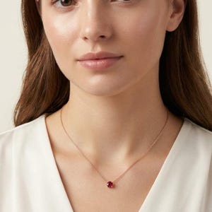 May include: A delicate rose gold chain necklace featuring a single, round, faceted red gemstone pendant set in a simple four-prong setting. The necklace rests against the collarbone, showcasing a minimalist design with a thin, polished metal chain.