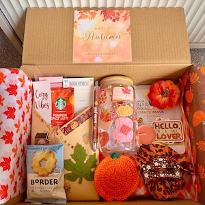 May include: A cardboard gift box filled with autumn-themed items including a glass tumbler with fall decals, a crochet pumpkin, a packet of Border shortbread rings, a Starbucks pumpkin spice latte mix, a snail collagen mask, and a Hello Fall sticker.