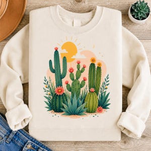 May include: A cream-coloured crewneck sweatshirt featuring a central graphic illustration of various green cacti and succulents with pink and yellow flowers, set against a backdrop of a yellow sun and soft orange clouds on a light-coloured fabric.