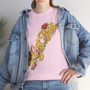 以下が含まれることがあります： 斜めに配置された黄色の刺繍テキストGot Thisと、赤いバラや小さなピンクの花の装飾的な蔓が描かれたライトピンクのTシャツ。金属製のボタンが付いた青いデニムジャケットと青いデニムジーンズを着用しています。