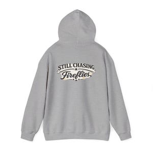 May include: A heather gray hooded sweatshirt featuring a graphic on the back with the text STILL CHASING Fireflies in black and white lettering, accented by small stars and a light, textured background design within the central graphic area.