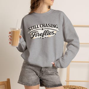 May include: A heather gray crewneck sweatshirt featuring the black text Still Chasing Fireflies in a stylized font with decorative stars and a subtle distressed background effect across the chest area, paired with gray denim shorts.