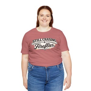 May include: A dusty rose short-sleeved t-shirt featuring a graphic print on the chest. The design includes the text "STILL CHASING Fireflies" in black and white lettering, accented with small stars and a distressed, light-colored background shape.
