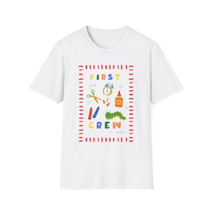 May include: A white short-sleeve t-shirt featuring a red dashed rectangular border. Inside, the text reads FIRST GRADE CREW. Illustrations include a blue alarm clock, green scissors, red push pins, a glue bottle, crayons, and a green caterpillar.