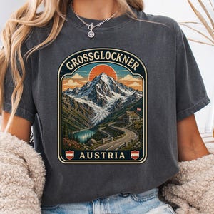 以下が含まれることがあります： オーストリアのグロースグロックナー山が描かれたグラフィックプリントのチャコールグレーの半袖Tシャツ。デザインには曲がりくねった道、建物、青い湖、夕日が描かれており、GrossglocknerとAustriaの文字がプリントされています。