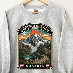 以下が含まれることがあります： オーストリアのグロースグロックナー山が中央にプリントされた、ヘザーグレーのクルーネックトレーナー。雪を頂いた山頂、曲がりくねった道路、建物、夕日が描かれたイラストで、GrossglocknerとAustriaの文字が添えられています。