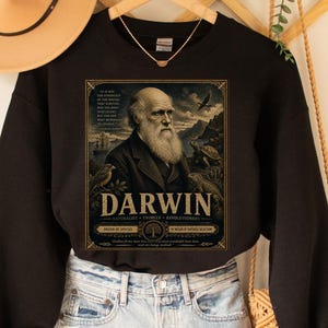 May include: A black long-sleeve sweatshirt featuring a graphic print of Charles Darwin with the text "DARWIN" and "NATURALIST - CYNICS - REVOLUTIONARY" alongside illustrations of a turtle, a bird, and a ship in a framed vintage-style design.